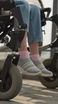Legs of little girl with multiple sclerosis sitting in wheelchair on walk with mother on street. Woman pushes medical transport with daughter closeup