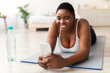 Happy plus size black woman using mobile phone on her break from domestic training, browsing web, looking up workout video online. Overweight African American lady checking cellphone after exercising