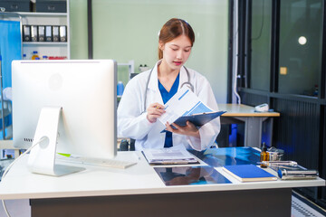 A young Asian female doctor, confidently dressed in a white uniform, sits at a computer desk in a...