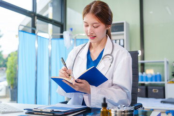 A young Asian female doctor, confidently dressed in a white uniform, sits at a computer desk in a...