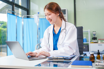 A young Asian female doctor, confidently dressed in a white uniform, sits at a computer desk in a hospital. She smiles while using a laptop, showcasing professionalism and dedication