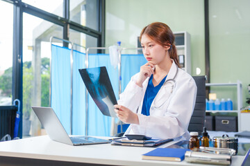 A young Asian female doctor, confidently dressed in a white uniform, sits at a computer desk in a hospital. She smiles while using a laptop, showcasing professionalism and dedication