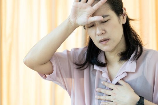 Asian woman experiencing symptoms of heart palpitations and dizziness,feel fatigued,holding chest,wiping sweat from her forehead with hand,hot flushes,weakness,overheating,dehydration,heat exhaustion