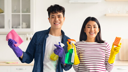 Smiling asian couple with supplies for cleaning house posing in kitchen, funny young korean man and...