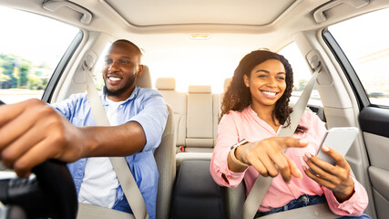 Portrait of smiling African American couple sitting inside luxury car, cheerful man holding steering wheel, woman with mobile phone pointing at the road, showing way, using digital map application