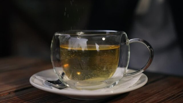 Person adding sugar into green tea in transparent cup at wooden table in housing. Moment captures human relaxation as sweetens tea in comfort of home. Human enjoys warm tea with sugar