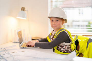 Young child in construction attire works on a laptop at a desk with blueprints during daylight hours