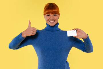 Cheerful woman shows a blank card while smiling and giving a thumbs up in front of a yellow backdrop