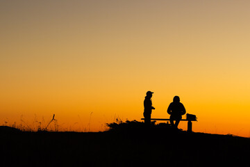 Friends sharing a quiet moment at sunset on a hilltop with vibrant orange sky. They enjoy a peaceful moment together, surrounded by nature's beauty.