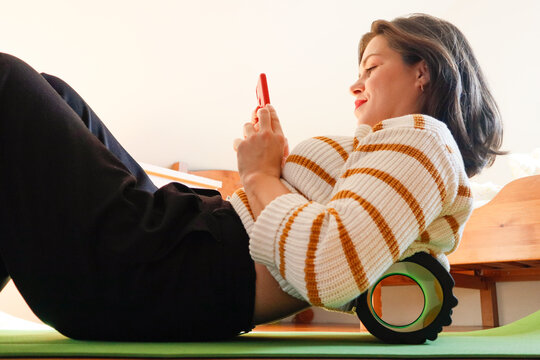 Woman massages her back with a roller at home with a phone in hands