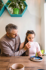 A father attentively helps his teenage daughter with Down syndrome eat a dessert at a cafe. He s gently assisting her, demonstrating care and support. 
