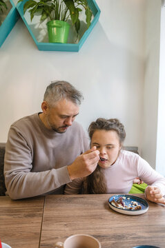 A father gently feeds his teenage daughter with Down syndrome a dessert at a cafe. He is showing loving care and support, highlighting the close bond between them. The image is warm and inviting.