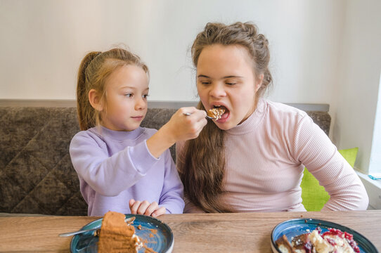 Two young girls are sharing a dessert at a cafe. One girl, who has Down syndrome, is being lovingly fed by her younger sister or friend. The scene is warm, cheerful