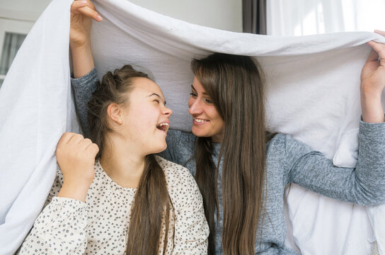 A young woman with Down syndrome and her mother are sharing a playful moment under a white sheet. They are laughing and looking at each other, showcasing a loving and supportive relationship. 