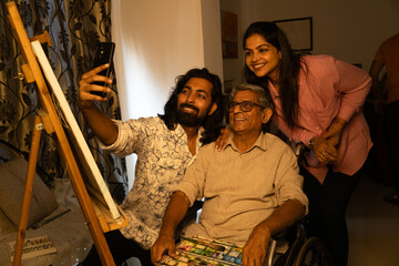 young indian couple taking selfie with senior disable father sitting on wheel chair with a painting brush and palette at home.