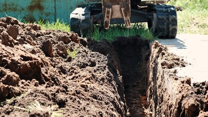 Fototapeta premium Mini excavator digs a narrow trench in soil for laying communications close-up. Mini bucket of crawler machine. Earthworks with heavy special equipment. Digging machinery works at construction site