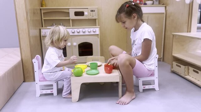 Children play in children's kitchen. Two girls in white clothes sit at small table and have fun, pretend to eat and drink. Children's games and activities at home.