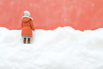 Child in an orange winter coat standing in deep snow against a coral wall. Concept of winter and youth.