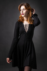 Red-haired gothic girl in a black dress on a gloomy background in the studio.