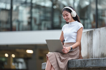 Young Asian woman is sitting outside a modern office building wearing headphones and listening to music on her mobile phone and laptop computer. Enjoy your working time. Relaxation concept.