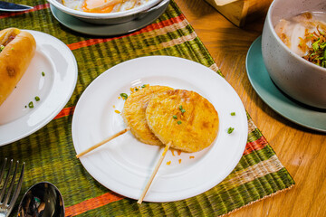 A plate features two circular snacks on sticks, garnished with herbs, set against a colorful, textured background, alongside other dishes.