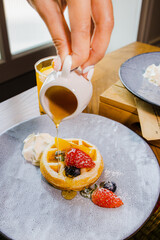 A hand pours syrup over a dessert featuring a waffle topped with fresh berries and cream, presented...