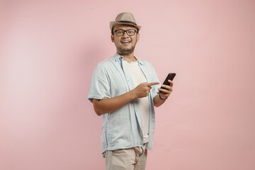 Smiling Asian man wearing hat and blue shirt holding and pointing to his smartphone.