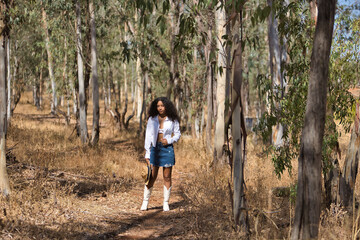 Obraz premium Latin woman, young, beautiful, dark-haired, with curly hair, with a white shirt and top, denim skirt and hat, walking alone and calmly through a eucalyptus forest. Concept peace, relaxation.