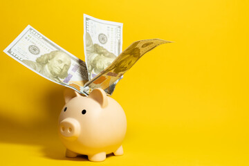 Side view of piggy bank filled with hundred dollar bills coming out of it on a yellow background with copy space, savings and economy concept © Answer 7