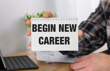 A person holding a spiral bound notebook with the words Begin New Career written on it. The notebook is on a desk with a keyboard and a laptop
