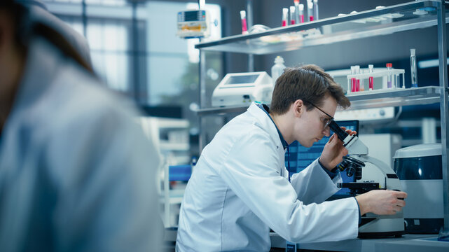 Molecular Biology Research Scientist Looking at a DNA Sample Under a Microscope in an Applied Science Laboratory. Portrait of a Young Lab Engineer in White Coat Working on Vaccine Products