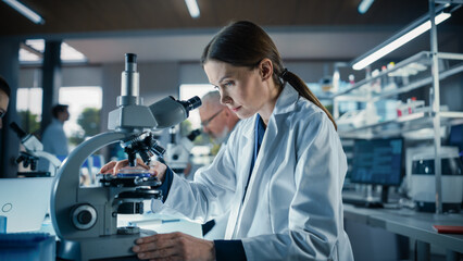 Female Bioengineer Looking at DNA Samples Under a Microscope in a Modern Applied Science Laboratory. Portrait of a Young Lab Engineer in White Coat Working with Colleagues