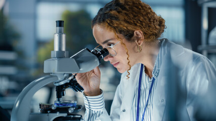 Hisplanic Female Bioengineer Looking at DNA Samples Under a Microscope in a Modern Applied Science Laboratory. Portrait of a Young Lab Engineer in White Coat Inventing New Medical Drugs