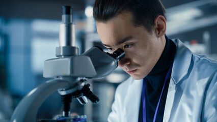 Medical Research and Development Laboratory: Young Caucasian Male Scientist Looking at a Biological Sample in a Petri Dish Under a Microscope in an Advanced Biotechnology Lab