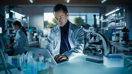 Portrait of a Young Graduate Student Working in a Laboratory, Using Laptop, Writing Down Research Data on a Computer. Multiethnic Biotechnology Specialist Using Modern Tools