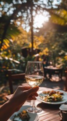 A hand holds a glass of white wine in a sunlit garden setting, capturing a relaxed outdoor dining experience.