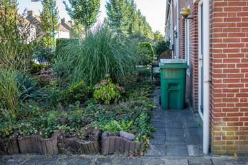 Residential garden with green trash can, brick house, and various plants.  Neatly landscaped with a...