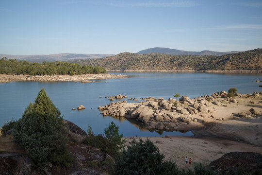 High-angle view of a reservoir with low water levels.  Rocky shoreline and hillsides are visible.  A couple walks along the dry, sandy beach.