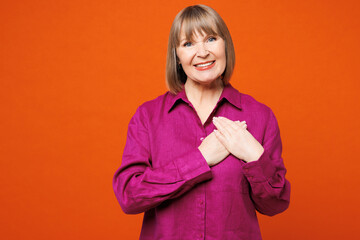 Fototapeta premium Elderly smiling happy blonde woman 50s years old she wears purple shirt casual clothes put folded hands on heart look camera isolated on plain red orange background studio portrait. Lifestyle concept.