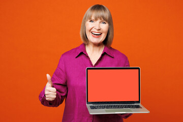 Elderly blonde woman 50s years old she wear purple shirt casual clothes hold use work on laptop pc computer with blank screen show thumb up isolated on plain red orange background. Lifestyle concept.