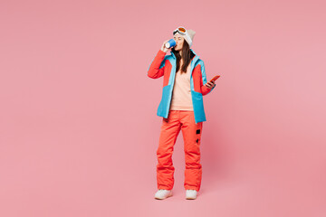 Full body skier young woman in warm blue padded jacket ski goggles mask spend weekend winter season in mountains hold use mobile cell phone drink tea isolated on plain pink background. Hobby concept.