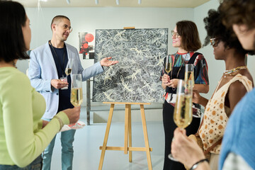 Young man with champagne explaining main idea of abstract painting while standing next to easel in front of public