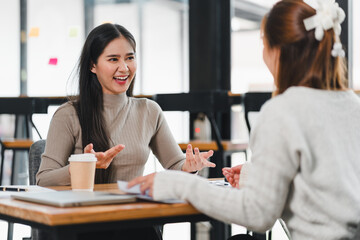 Two women having friendly conversation at table in modern office space, with one holding coffee cup...