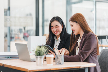 Two businesswomen in formal attire working together at desk with laptop, tablet, and documents in modern office. They are smiling and collaborating effectively