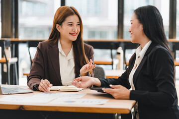 Two businesswomen in formal attire discussing work at desk with documents and laptop, smiling and engaging in professional conversation in modern office