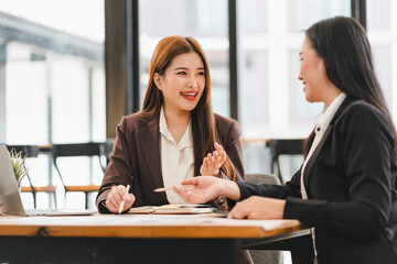 Two professional women in business attire having discussion at modern office table, with laptop and documents, showcasing collaboration and teamwork