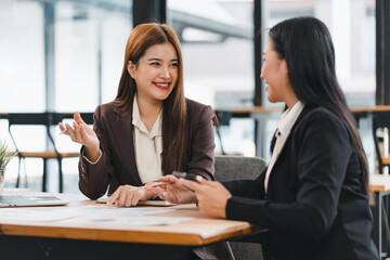 Two businesswomen in formal attire having cheerful discussion at modern office table, with documents and laptop in front of them, showcasing teamwork and collaboration