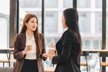 Two professional women in business attire having friendly conversation while holding coffee cups in modern office with large windows