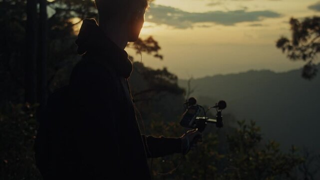 A young man holds a microphone to record his surroundings against the backdrop of a mountain sunset