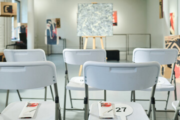 Focus on several rows of white plastic chairs with brochures and bidding paddles standing in front of abstract painting for sale